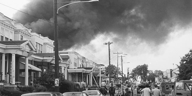 Smoke billows over rowhouses in the West Philadelphia after the police bombed the home of the radical African American organization MOVE during a standoff. Police on horseback and emergency vehicles block off a street as residents walk towards the scene.