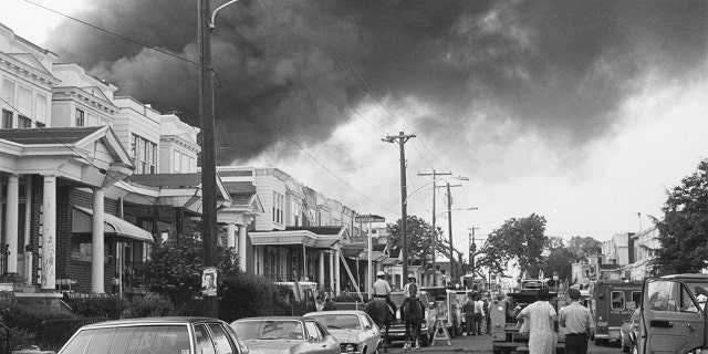 Smoke billows over rowhouses in the West Philadelphia after the police bombed the home of the radical African American organization MOVE during a standoff. Police on horseback and emergency vehicles block off a street as residents walk towards the scene.