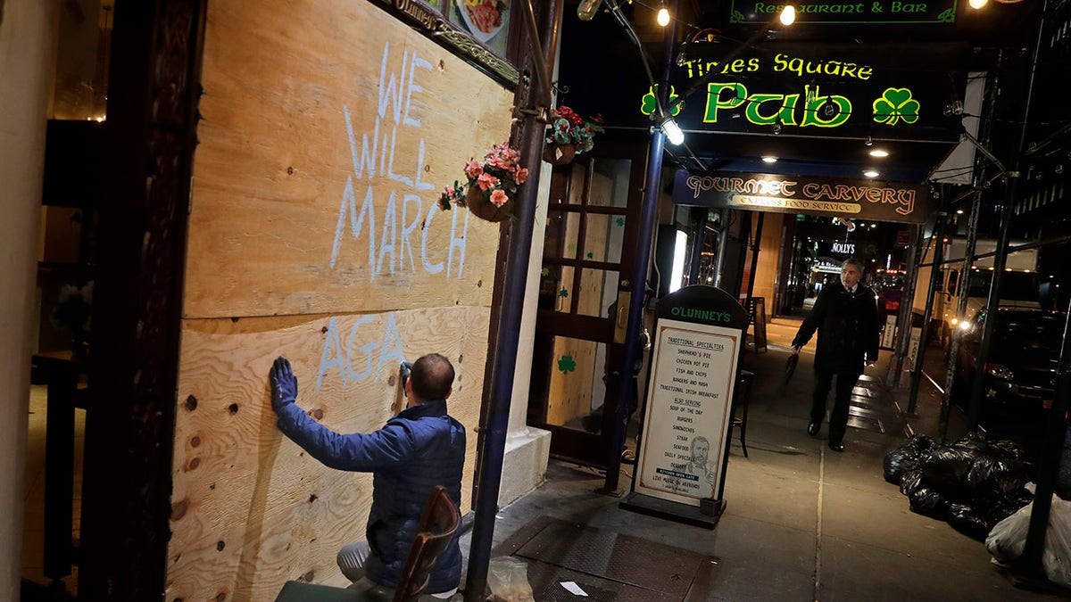 FILE - The manager of O'Lunney's Times Square Pub, John Galgey, writes "We Will March Again!" on the plywood he used to board up the large glass windows of the pub in New York, Monday, March 16, 2020. More than a year after coronavirus shutdowns sent "the city that never sleeps" into a fitful slumber, New York could soon be wide awake again. Vaccinated New Yorkers can shed their masks in most situations Wednesday, and restaurants, shops, gyms and many other businesses can go back to full occupancy if all patrons are inoculated. (AP Photo/Seth Wenig, File)