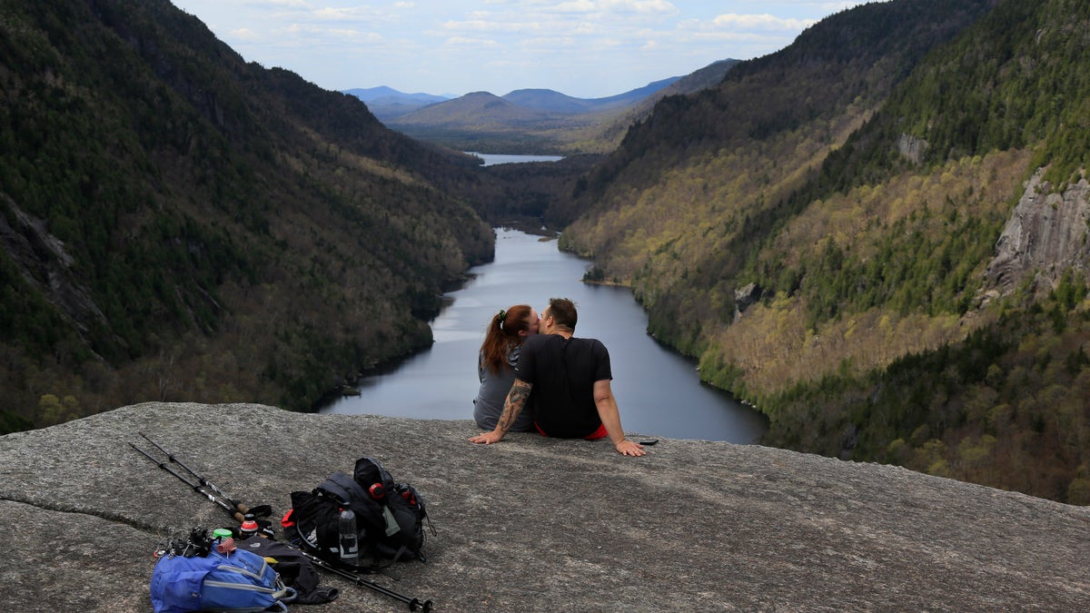 Sidney Gleason, left, and Joe Gorsuch, both of Syracuse, kiss while taking in the view of Lower Ausable Lake at Indian Head summit inside the Adirondack Mountain Reserve, Saturday, May 15, 2021, near St. Huberts, N.Y. A free reservation system went online recently to control the growing number of visitors packing the parking lot and tramping on the trails through the private land of the Adirondack Mountain Reserve. The increasingly common requirements, in effect from Maui to Maine, offer a trade-off to visitors, sacrificing spontaneity and ease of access for benefits like guaranteed parking spots and more elbow room in the woods. (AP Photo/Julie Jacobson)