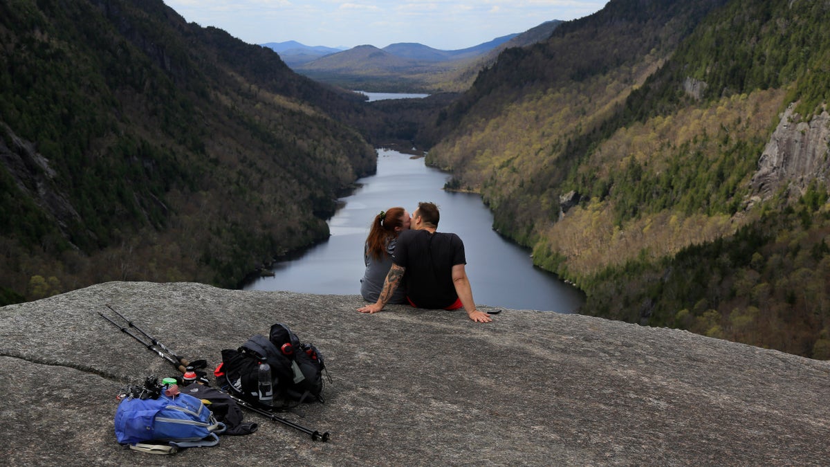 Sidney Gleason, left, and Joe Gorsuch, both of Syracuse, kiss while taking in the view of Lower Ausable Lake at Indian Head summit inside the Adirondack Mountain Reserve, Saturday, May 15, 2021, near St. Huberts, N.Y. A free reservation system went online recently to control the growing number of visitors packing the parking lot and tramping on the trails through the private land of the Adirondack Mountain Reserve. The increasingly common requirements, in effect from Maui to Maine, offer a trade-off to visitors, sacrificing spontaneity and ease of access for benefits like guaranteed parking spots and more elbow room in the woods. (AP Photo/Julie Jacobson)