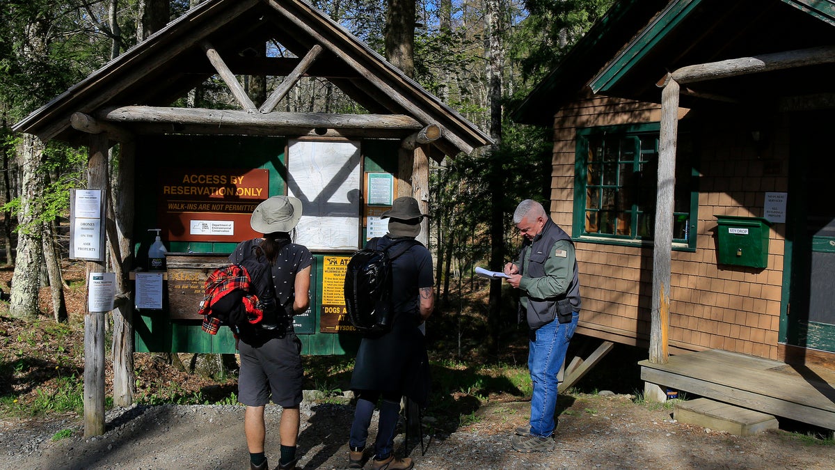 Adirondack Mountain Reserve Ranger Mike Ryan, right, checks in two hikers at the trailhead of the reserve, Saturday, May 15, 202, in St. Huberts, N.Y. A free reservation system went online recently to control the growing number of visitors packing the parking lot and tramping on the trails through the private land of the Adirondack Mountain Reserve. The increasingly common requirements, in effect from Maui to Maine, offer a trade-off to visitors, sacrificing spontaneity and ease of access for benefits like guaranteed parking spots and more elbow room in the woods. (AP Photo/Julie Jacobson)