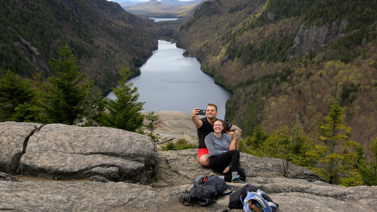 Sidney Gleason, right, and Joe Gorsuch, both of Syracuse, take a selfie with Lower Ausable Lake in the background, while taking a break at Indian Head summit inside the Adirondack Mountain Reserve, Saturday, May 15, 2021, near St. Huberts, N.Y. A free reservation system went online recently to control the growing number of visitors packing the parking lot and tramping on the trails through the private land of the Adirondack Mountain Reserve. The increasingly common requirements, in effect from Maui to Maine, offer a trade-off to visitors, sacrificing spontaneity and ease of access for benefits like guaranteed parking spots and more elbow room in the woods. (AP Photo/Julie Jacobson)