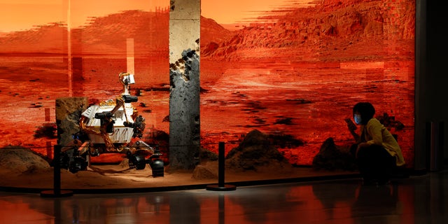 A woman wearing a mask talks on her phone near an exhibition depicting a rover on Mars in Beijing on Friday, May 14, 2021. China says its Mars probe and accompanying rover landed on the red planet. (AP Photo/Ng Han Guan)