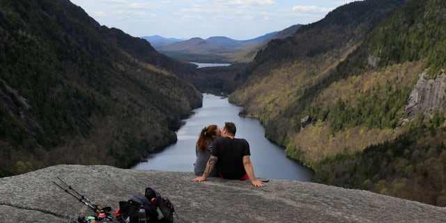 Sidney Gleason, left, and Joe Gorsuch, both of Syracuse, kiss while taking in the view of Lower Ausable Lake at Indian Head summit inside the Adirondack Mountain Reserve, Saturday, May 15, 2021, near St. Huberts, N.Y. A free reservation system went online recently to control the growing number of visitors packing the parking lot and tramping on the trails through the private land of the Adirondack Mountain Reserve. The increasingly common requirements, in effect from Maui to Maine, offer a trade-off to visitors, sacrificing spontaneity and ease of access for benefits like guaranteed parking spots and more elbow room in the woods. (AP Photo/Julie Jacobson)