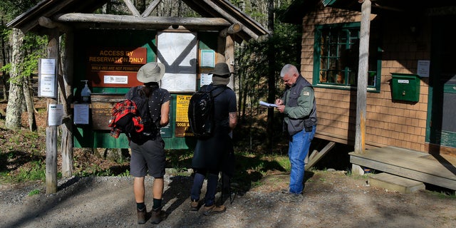 Adirondack Mountain Reserve Ranger Mike Ryan, right, checks in two hikers at the trailhead of the reserve, Saturday, May 15, 202, in St. Huberts, N.Y. A free reservation system went online recently to control the growing number of visitors packing the parking lot and tramping on the trails through the private land of the Adirondack Mountain Reserve. The increasingly common requirements, in effect from Maui to Maine, offer a trade-off to visitors, sacrificing spontaneity and ease of access for benefits like guaranteed parking spots and more elbow room in the woods. (AP Photo/Julie Jacobson)