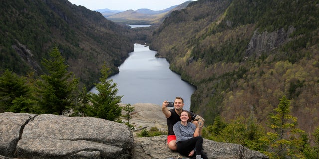 Sidney Gleason, right, and Joe Gorsuch, both of Syracuse, take a selfie with Lower Ausable Lake in the background, while taking a break at Indian Head summit inside the Adirondack Mountain Reserve, Saturday, May 15, 2021, near St. Huberts, N.Y. A free reservation system went online recently to control the growing number of visitors packing the parking lot and tramping on the trails through the private land of the Adirondack Mountain Reserve. The increasingly common requirements, in effect from Maui to Maine, offer a trade-off to visitors, sacrificing spontaneity and ease of access for benefits like guaranteed parking spots and more elbow room in the woods. (AP Photo/Julie Jacobson)