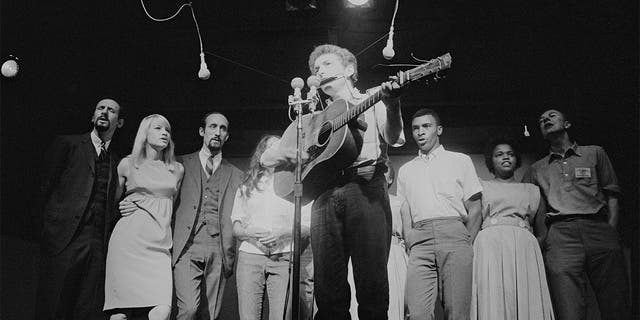 American musician Bob Dylan plays acoustic guitar and harmonica during a performance at the Newport Folk Festival, Newport, Rhode Island, July 1963. Among those behind him are, from left, Peter Yarrow, Mary Travers (1936 - 2009, Paul Stookey, Joan Baez (partially obscured), two unidentified people, Charles Neblett, Rutha Harris, and Pete Seeger (1919 - 2014).