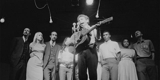 American musician Bob Dylan plays acoustic guitar and harmonica during a performance at the Newport Folk Festival, Newport, Rhode Island, July 1963. Among those behind him are, from left, Peter Yarrow, Mary Travers (1936 - 2009, Paul Stookey, Joan Baez (partially obscured), two unidentified people, Charles Neblett, Rutha Harris, and Pete Seeger (1919 - 2014).