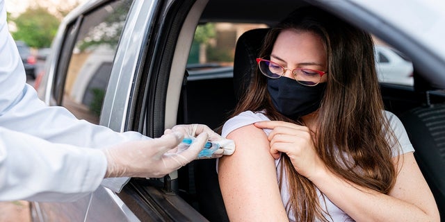 A woman with a mask on is shown receiving a COVID vaccination from her car. 