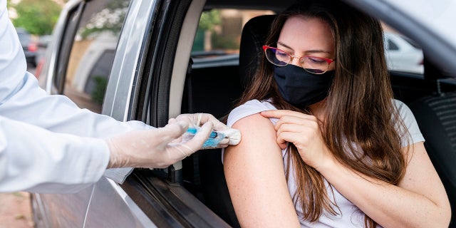 A woman with a mask on is shown receiving a COVID vaccination from her car.