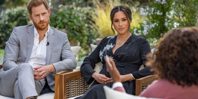 Prince Harry in a grey suit and white shirt sits next to wife Meghan Markle in a blue dress with light blue detail, across from the back of Oprah Winfrey doing an interview