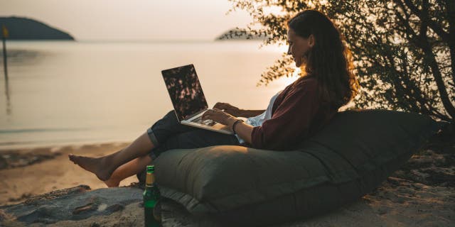 A woman sits outside with her laptop on the summer beach at sunset.