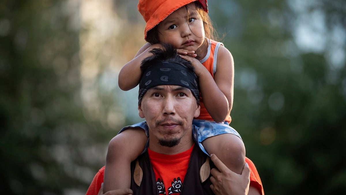 Cowichan Tribe member Benny George holds his child Bowie, 3, on his shoulders as they listen during a ceremony and vigil for the 215 children whose remains were found buried at the former Kamloops Indian Residential School, in Vancouver, British Columbia, on National Indigenous Peoples Day, Monday, June 21, 2021. (Darryl Dyck/The Canadian Press via AP)