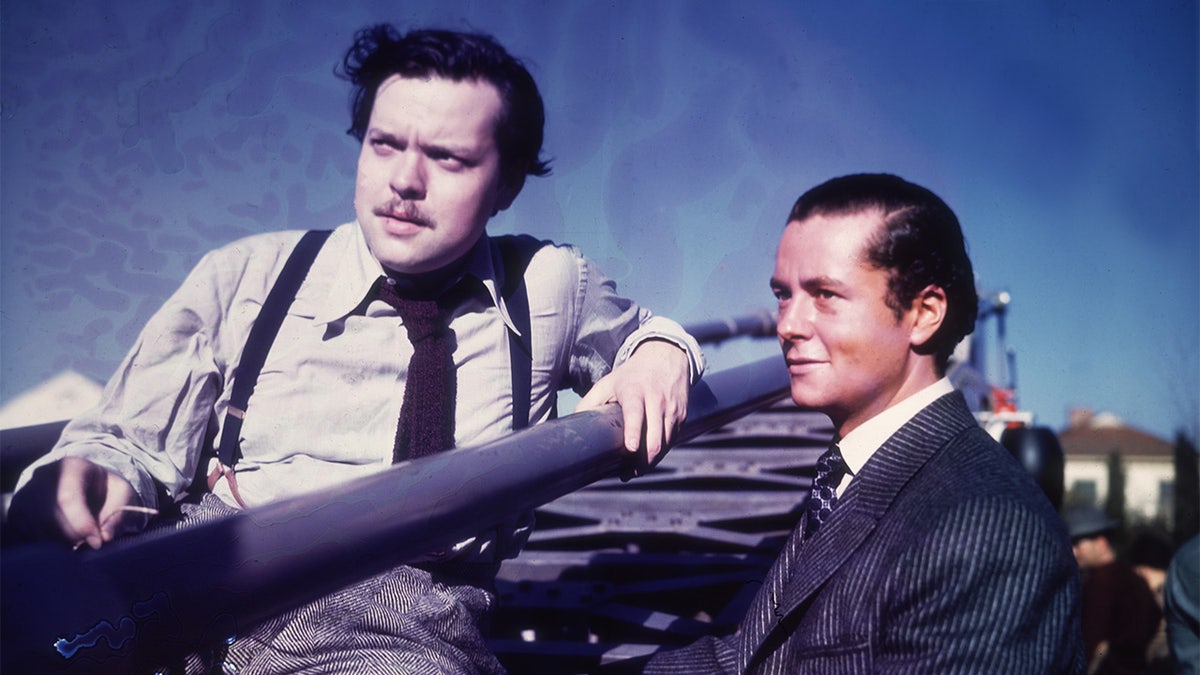 1942: American actor, producer and director Orson Welles sits in a stadium while American actor Tim Holt stands next to him outdoors on the set of Welles' film, 'The Magnificent Ambersons'. Holt is dressed in period costume. (Photo by Hulton Archive/Getty Images)