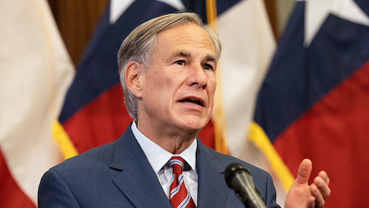 Gov. Greg Abbott seen speaking at press conference wearing striped red tie and standing in front of flags
