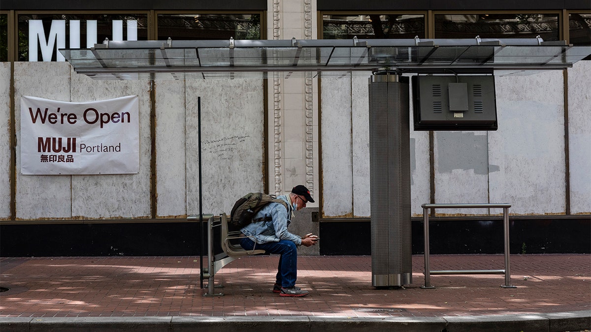 A man sits at a bus stop as wooden panels barricade shops because of on-going protests on Saturday, June 5, 2021 in downtown Portland, Ore. City officials insist Portland is resilient as they launch a revitalization plan — in the form of citywide cleanups of protest damage, aggressive encampment removals, increased homeless services and police reform — to repair its reputation.