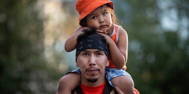 Cowichan Tribe member Benny George holds his child Bowie, 3, on his shoulders as they listen during a ceremony and vigil for the 215 children whose remains were found buried at the former Kamloops Indian Residential School, in Vancouver, British Columbia, on National Indigenous Peoples Day, Monday, June 21, 2021. (Darryl Dyck/The Canadian Press via AP)