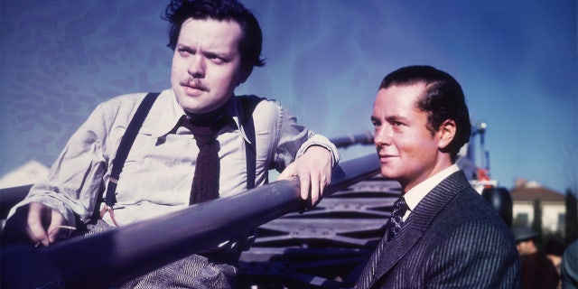 American actor, producer and director Orson Welles produced a definitive work about aliens called ‘The War of the Worlds.’ Here he sits in a stadium while American actor Tim Holt stands next to him outdoors on the set of Welles' film, 'The Magnificent Ambersons.' Holt is dressed in period costume. Â