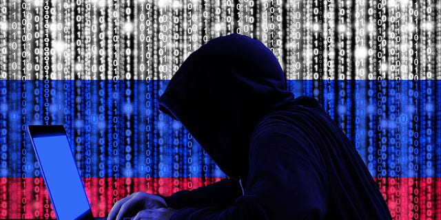 Hacker in a dark hoodie sitting in front of laptop with digital Russian flag and cyber security concept on binary streams background