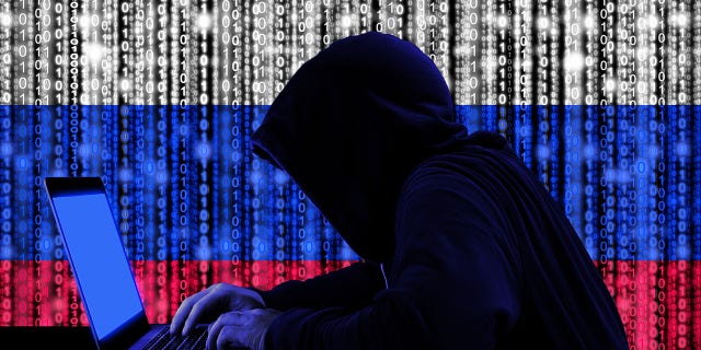 Hacker in a dark hoodie sitting in front of laptop with digital Russian flag and cyber security concept on binary streams background