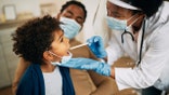 Family doctor examining throat of a small black boy while visiting him at home during coronavirus pandemic.