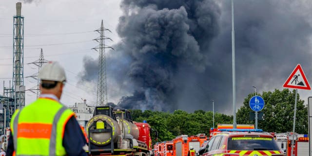 Emergency vehicles of the fire brigade, rescue services and police stand not far from an access road to the Chempark over which a dark cloud of smoke is rising in Leverkusen, Germany, Tuesday, July 27, 2021.