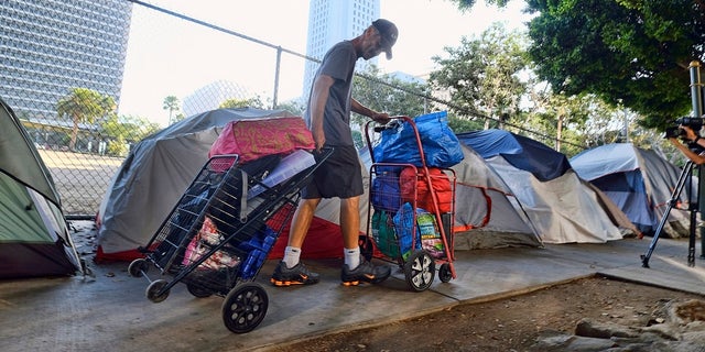 FILE - In this July 1, 2019 file photo, a homeless man moves his belongings from a street near Los Angeles City Hall, background, as crews prepared to clean the area. The Los Angeles City Council has passed a sweeping anti-camping measure to remove widespread homeless encampments that have become an eyesore across the city. Among other limits, the ordinance that passed 13-2 would ban sitting, lying, sleeping or storing personal property that blocks sidewalks, streets and bike lanes or near driveways, fire hydrants, schools, day care centers, libraries, homeless shelters and parks. (AP Photo/Richard Vogel, File)