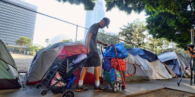 In this July 1, 2019 file photo, a homeless man moves his belongings from a street near Los Angeles City Hall, background, as crews prepared to clean the area. The Los Angeles City Council has passed a sweeping anti-camping measure to remove widespread homeless encampments that have become an eyesore across the city. (AP Photo/Richard Vogel, File)