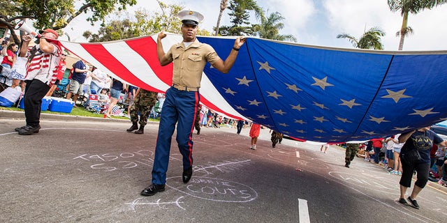 یک تفنگدار آمریکایی و رژه‌بازان، پرچم بزرگ آمریکا را در رژه چهارم جولای کورونادو در 3 ژوئیه 2021 در کورونادو، کالیفرنیا حمل می‌کنند.