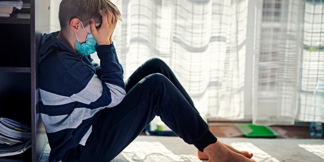 Depressed little boy sitting by the window, wearing surgical mask