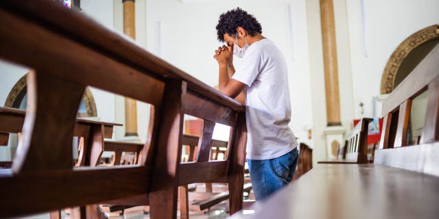 Young man, wearing a white protective mask, inside the church, praying in times of coronavirus
