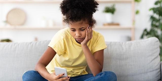 فتاة مراهقة أمريكية من أصل أفريقي تشعر بالملل وهي تستخدم الهاتف الذكي وتتصفح الإنترنت جالسة على الأريكة في المنزل.  طفولة مملة حديثة