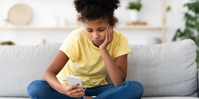 Bored African American Teen Girl Using Smartphone Browsing Internet Sitting On Sofa At Home. Modern Boring Childhood