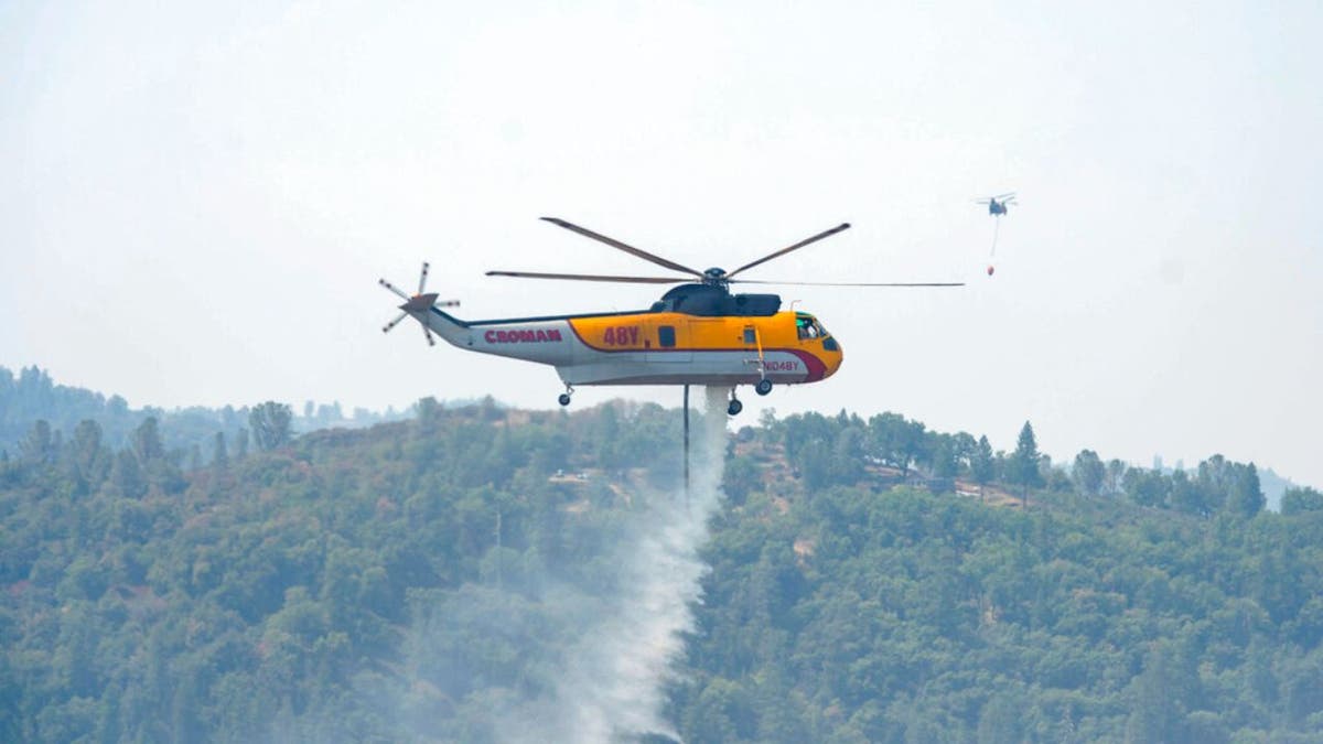 A Croman Sikorsky S-61A helicopter drops water on the south end of the Caldor Fire near Grizzly Flat Road west of Grizzly Flats, Calif., on Monday, Aug. 23, 2021, as a second helicopter flies over a nearby home as it departs in the distance to refill its water bucket. 