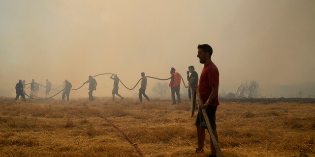 A firefighter tries to extinguish a fire as volunteers hold the water hose in Agios Stefanos, in northern Athens, Greece, Friday, Aug. 6, 2021. Thousands of people have fled wildfires burning out of control in Greece and Turkey, including a major blaze just north of the Greek capital of Athens that left one person dead. (AP Photo/Thanassis Stavrakis)