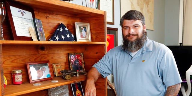 In this Aug. 11, 2021, photo William Bee poses for a photo next to a bookshelf that displays items from his families military service including his Purple Heart at his home in Jacksonville, N.C. (AP Photo/Karl B DeBlaker)