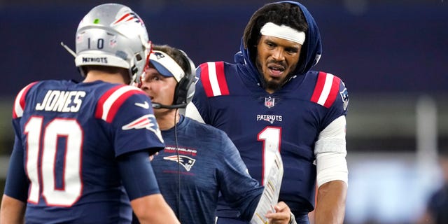 New England Patriots quarterback Cam Newton (1) looks on as offensive coordinator Josh McDaniels talks with quarterback Mac Jones during the first half of the team's preseason game against Washington, Aug. 12, 2021, in Foxborough, Massachusetts.