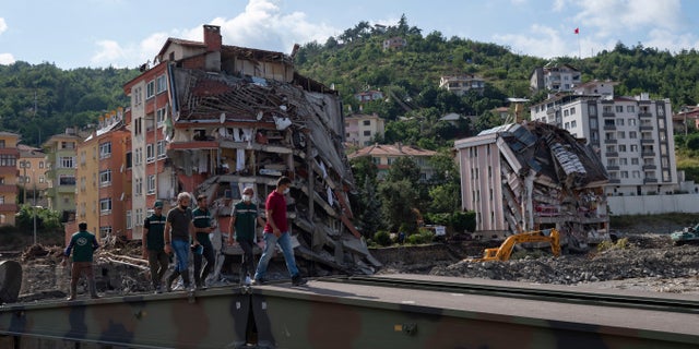 Cilvēki staigā pa pagaidu tiltu, ko militāristi uzcēla Bozkurtas pilsētā Turcijas Kastamonu provincē svētdien, 2021. gada 15. augustā, pēc plūdiem.  Turcija ir nosūtījusi kuģus, lai palīdzētu evakuēt cilvēkus un transportlīdzekļus no plūdos skartās Melnās jūras ziemeļu pilsētas, jo svētdien notikušās katastrofas upuru skaits pieauga līdz vismaz 62 un daudzi citi joprojām ir pazuduši.  (AP fotoattēls)