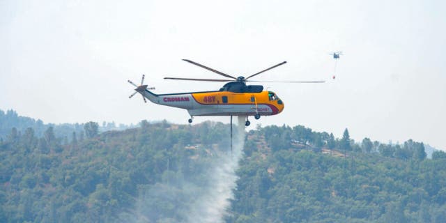 A Croman Sikorsky S-61A helicopter drops water on the south end of the Caldor Fire near Grizzly Flat Road west of Grizzly Flats, Calif., on Monday, Aug. 23, 2021, as a second helicopter flies over a nearby home as it departs in the distance to refill its water bucket.