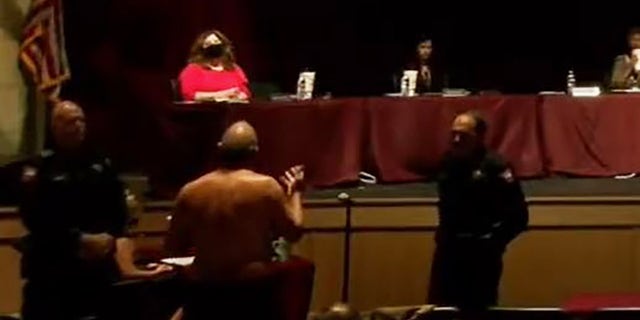 A shirtless James Akers, back to camera, addresses the school board in Dripping Springs, Texas, earlier this week. (School board video image)