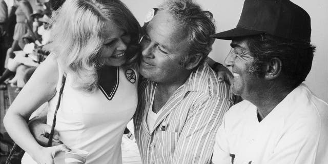 American actors Sally Struthers, Carroll O'Connor (1924-2001) and Dean Martin (1917-1995) chat in the dugout during a celebrity baseball game. Martin wears a Los Angeles Dodgers uniform and Struthers wears a minidress with the team logo. 