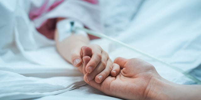 A child in a hospital bed holds their parents hand.