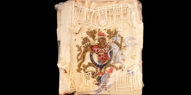 The wedding cake slice has a coat-of-arms design colored in red, silver, gold and blue. It also features a small silver horseshoe and leaf spray with white decorative icing. (Dominic Winter Auctioneers via AP)