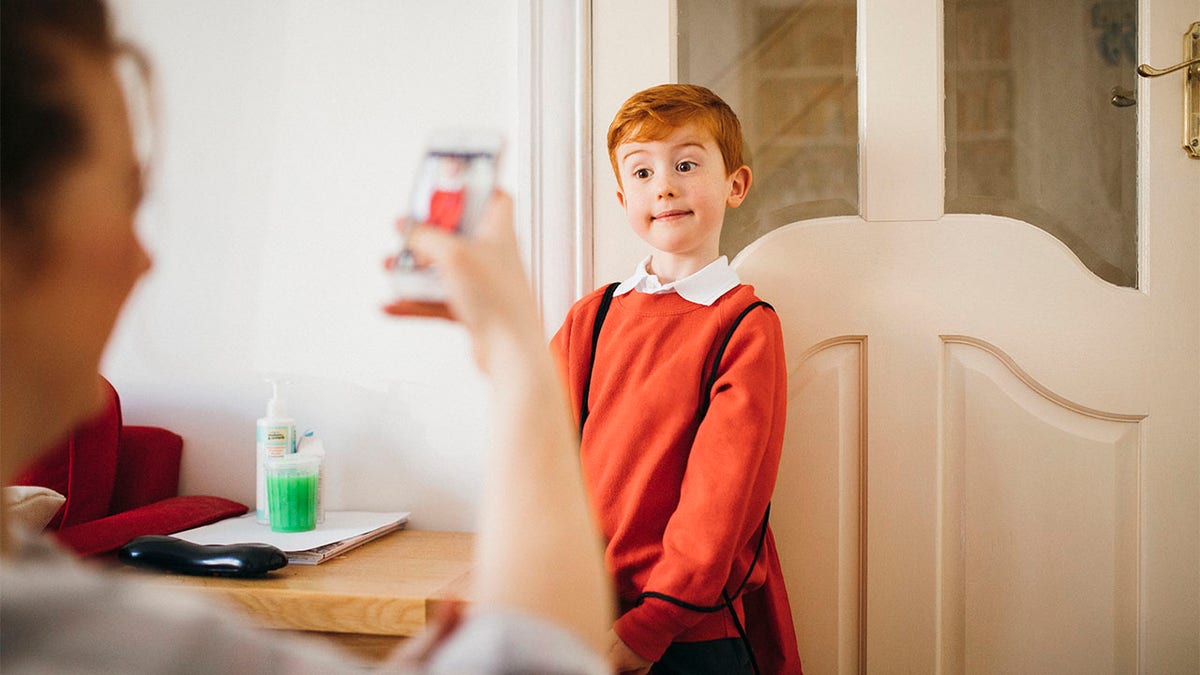 Little boy dressed for his first day back to school, having his photo taken by his Mother on her smartphone.