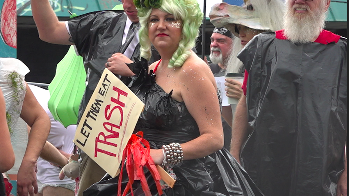 A woman holds a "Let them eat trash" sign while wearing a garbage bag for a dress at an organized 'trash parade' protest.