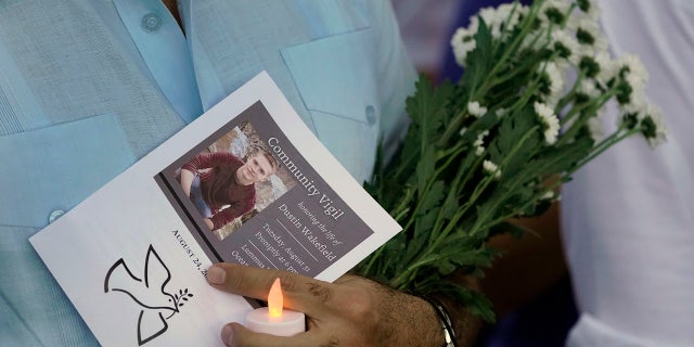 An attendee at a community vigil honoring the life of Dustin Wakefield, holds a program, flowers and a battery-powered candle, Tuesday, Aug. 31, 2021, in Miami Beach, Fla. Wakefield, 21, was shot and killed last week by gunman who walked into a Miami Beach restaurant and fatally shot Wakefield who was vacationing from Castle Rock, Colo. (AP Photo/Wilfredo Lee)