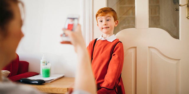 Little boy dressed for his first day back to school, having his photo taken by his Mother on her smartphone.