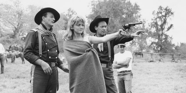 John Wayne and William Holden assist Constance Towers in aiming a revolver on the set.