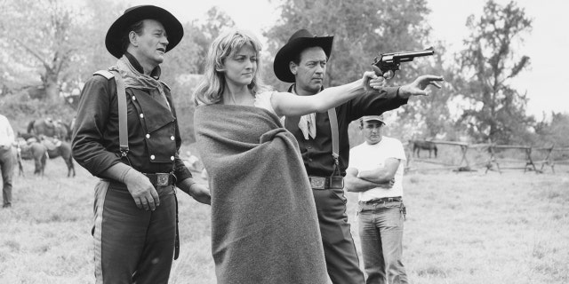 John Wayne and William Holden assist Constance Towers in aiming a revolver on the set.